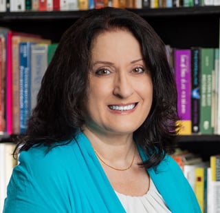 Woman with long dark hair smiling at camera, wearing turquoise blazer over white top, with bookshelves in background
