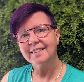 Smiling person with purple hair and glasses wearing a turquoise shirt, posed against a green foliage background