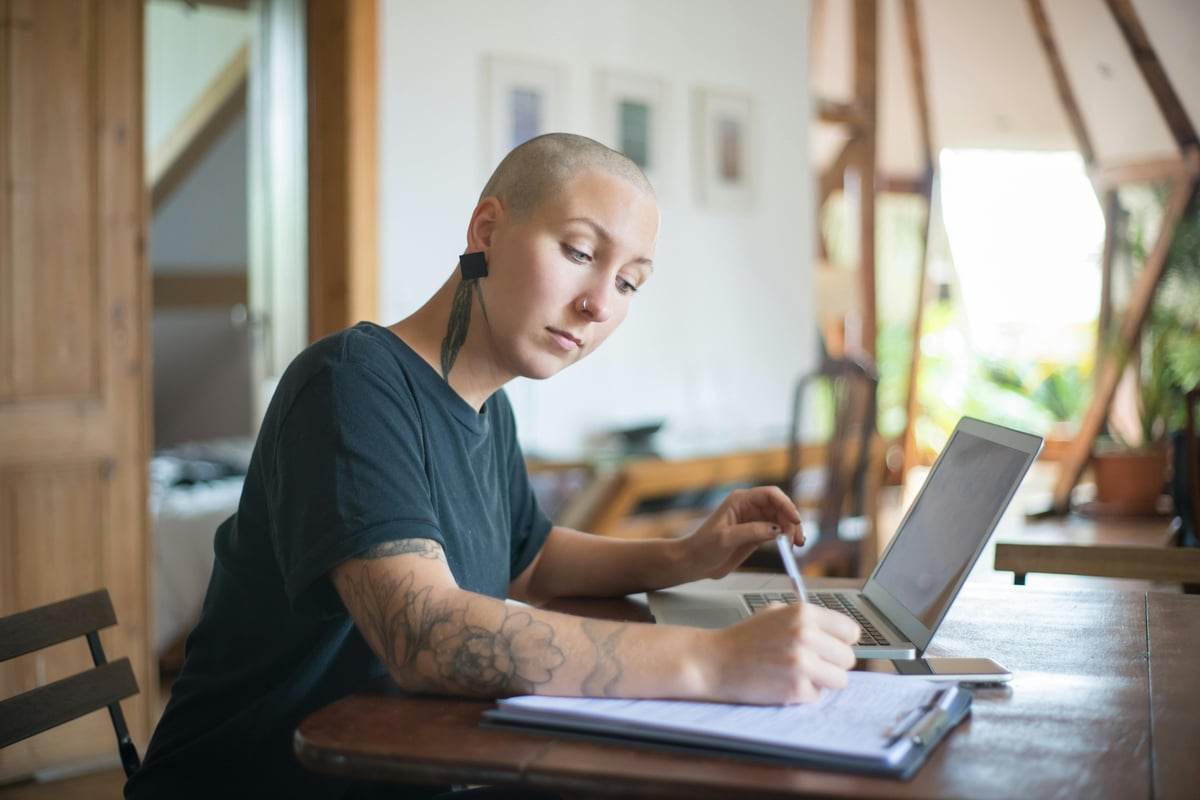 Person with tattoo working on laptop at wooden desk with notebook and pen in bright home office