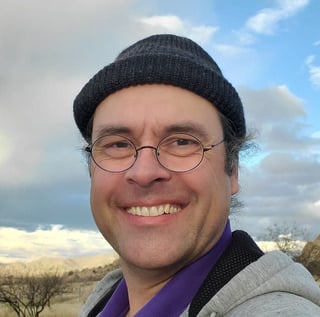 Portrait of a smiling man wearing glasses, a black beanie, and purple jacket outdoors with a cloudy sky and landscape background