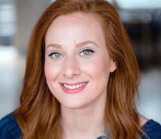 Professional headshot of a woman with reddish-brown hair and blue eyes smiling at the camera wearing a dark blue top