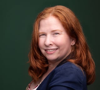 Woman with long reddish-brown hair smiling at camera against green background, wearing navy blazer