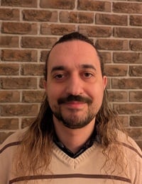 Man with long dark hair and mustache wearing a beige shirt against a brick wall background