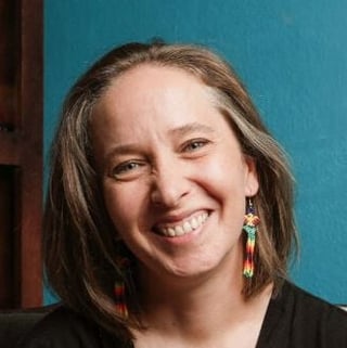 Portrait of a smiling woman with long brown hair wearing a black shirt and beaded earrings against a blue background