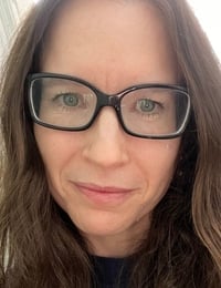 Woman with long brown hair and black-framed glasses looking at camera in a close-up portrait